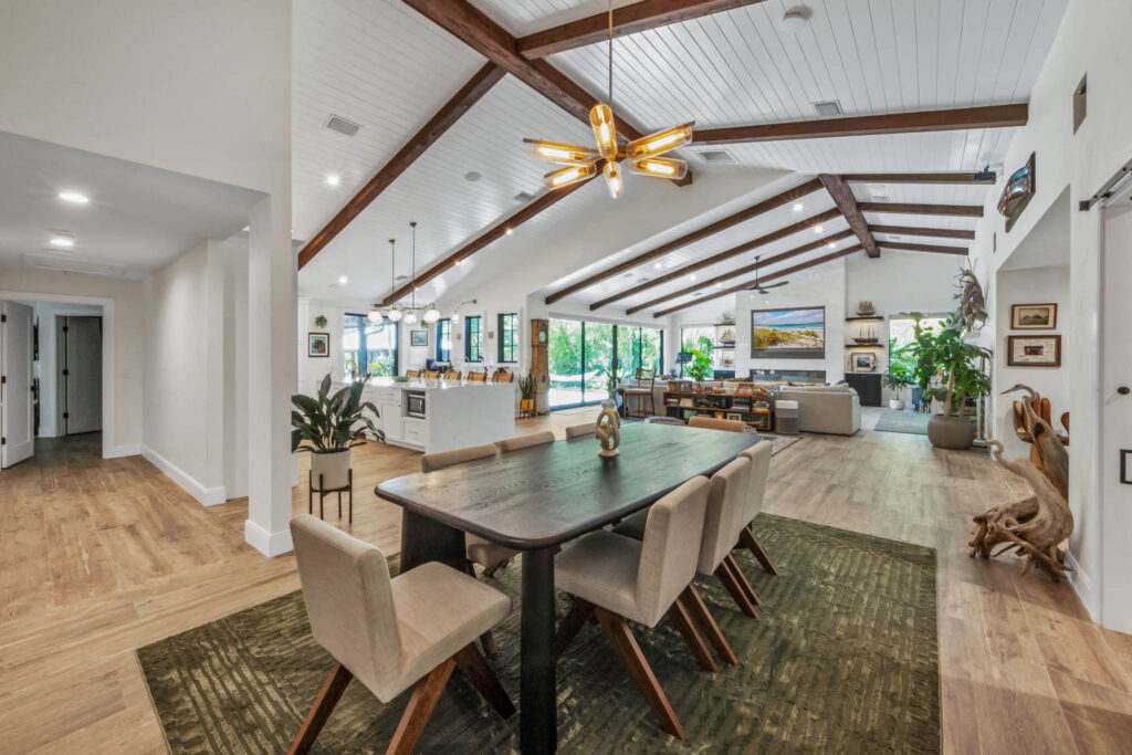 Eason Builders interior home design: living and dining area with a vaulted ceiling, exposed beams, a modern chandelier, a wooden table with six chairs, and wood flooring.